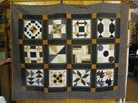 Full view of the entire quilt hanging showing the 12 patchwork blocks arranged in rows and columns with dark borders and burnt orange corner squares.