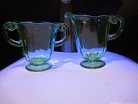 Both uranium glass pieces glowing under UV light, showing characteristic bright green fluorescence. Cream pitcher to the right with a handle and spout. Sugar bowl to the left with two handles.