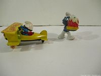 Two vintage Smurfs figurines displayed on white background, one driving yellow biplane 'Aero-Smurfs', the other carrying cake
