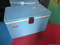Front and side view of vintage Coleman cooler showing light blue metal body, cream trim, red logo, and metal latch on hinged lid