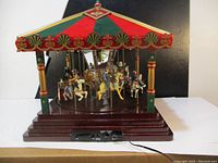 Front view of the Christmas carousel music box showing multicolored carousel horses and figures under red and green canopy with gold accents. Control panel and power cord in front base visible.