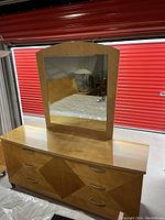 Photo showing a large wooden dresser with six drawers. The dresser features a geometric pattern on drawer fronts and a large attached mirror with a wood frame. The finish is tan/golden color and the dresser measures approximately 70 inches wide and 21 inches deep.