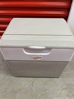 Front and top view of Koolatron cooler showing gray body with white top and handle, marked with Koolatron logo.