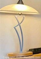 Side table lamp lit up showing the opaque round glass shade and the brushed silver-colored stem with geometric design on top of an oval wooden base