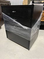 Side and front view of the black wooden office cabinet showing three drawers wrapped partially in clear plastic wrap, highlighting the cabinet's shape, drawer size differences, and wear on the paint.