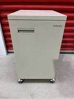 Front view of beige Toshiba steel office cabinet with caster wheels and Toshiba logo on door.