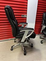 Side profile photo showing black bonded leather chair with padded armrest and caster wheel base, set against a red and white storage background.