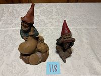 Two gnome figurines displayed on a patterned cloth surface. Left gnome wears an orange hat, holds a bottle, and is surrounded by seashells. Right gnome wears a red hat and plays a harmonica, sitting with bent knees.