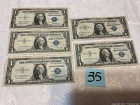 Five one-dollar silver certificates laid out on patterned white fabric, showing front of notes with blue seals and George Washington portraits.