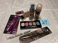 Lot contents including various unopened makeup items arranged on a surface, showing mascara, concealer, lipstick, foundation, eyeshadow palette, powder, eyeliners, and brow pencil.