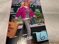 Barbie doll dressed in gray outfit with pink bomber jacket, carrying a 'big brown bag' shopping bag in front of Bloomingdale's illustrated background on the box.
