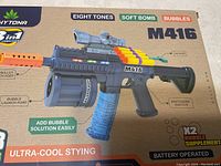 Photo of the front side of the toy gun box showing the M416 toy gun with labels pointing to parts like soft bullet launch port, bubble launch port, switching switch, bubble supplement, and highlighting features such as eight tones and battery operation.
