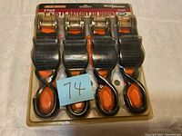 Photo shows four ratchet tie downs in original packaging with orange straps, metal ratchets, and black cushioned handles with orange grips.