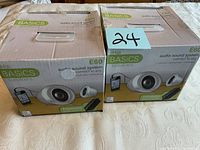 Two iHip Basics E60 speaker boxes placed on a white quilted fabric surface, showing front and side packaging with product images and features.