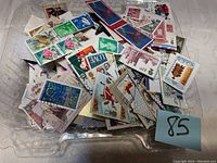 Top view of the plastic container filled with various loose used foreign stamps showing designs of flowers, buildings, ships, and other subjects.