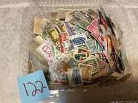 Photo showing assorted postage stamps in a clear plastic container, mostly used with various colors and countries represented.