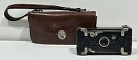 Photo of folded black Ensignette folding camera with silver metal accents next to a brown leather carrying case with clasp and handle strap.