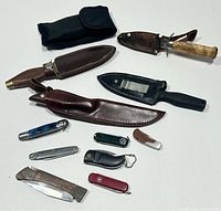 Full layout of twelve knives: top row nylon pouch and black folding liner-lock, middle row five fixed blades with various sheaths, bottom row six folding slipjoint knives and one unsheathed fixed blade