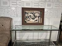 Framed mixed media artwork on paper displayed on a glass console table against a patterned wall.