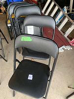 Six black folding chairs stacked in garage showing padded seats and backrests with metal frames.