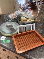 Multiple glass pie plates with lids, clear glass and stainless steel mixing bowls, copper wire drying rack, and assorted kitchen utensils on countertop.