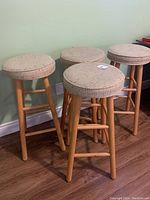 Four wooden leg bar stools with round beige upholstered seats arranged together against a wall on wood flooring.