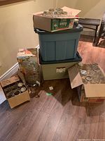 View of stacked plastic totes with boxes of canning jars around and a glass drink dispenser on metal stand.