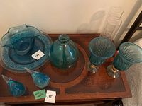 Photo showing full assortment of teal blue glassware on wooden table including platter, bowl, vase, goblets, bird figurines, and two clear glasses.