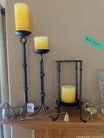 Photo of tall and short black metal candle holders each with yellow candles, brass decorative dachshund, elephants, and bell-shaped item on wooden surface.