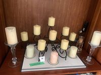 Front view showing black metal candelabra with candles and two clear glass candle holders each with a pillar candle, displayed on a wooden surface against a wood panel wall.