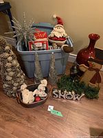 Lot arranged with most items around and inside a large gray plastic tote. Visible are the silver star ornament, Santa figurine sitting on the tote, two silver cone trees, pom-pom snowmen, snowman wall hanging, lantern, red vase, and green LED garland.