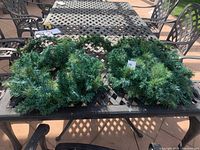 Three items including a Christmas garland with outdoor lights and two green artificial pine wreaths placed on a patio table outdoors under natural light.