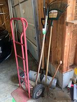 Photo showing red dolly with two wheels standing upright with shovels and rake leaning against the wall in garage setup.