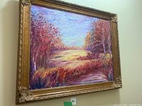 Frontal photo of a colorful landscape canvas painting in a gold ornate frame hung on a wall.