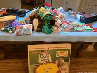 Wide view of the lot showing stuffed animal characters, vintage board games, children's books, and clothing items spread out on a table.