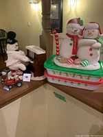 Photo showing fabric snowman couple, holiday containers stacked below, toy tractor with Santa and small snowman figure on acorn base, all displayed on wooden ledge.
