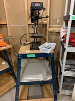 Jobmate drill press mounted on a blue metal stand with wheels, white basket on the stand shelf holding drill bits and materials below shelf