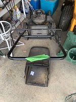 Lawnmower positioned in garage with grass catcher bag detached and placed behind, foldable handlebar in front with a green paper label.