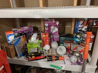 Photo showing assorted light bulbs of various colors, sizes, types, and packaging displayed on a white shelf including LED and incandescent bulbs and orange light bulb changer tool.