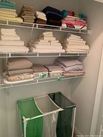 Closet shelves filled with stacked linens including light blue, beige, brown, white, grey, and pink folded fabrics plus three laundry hampers at bottom