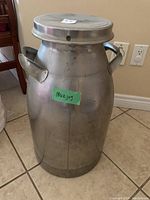 Front view of large silver metal milk jug with two handles and a lid. Green sticker label visible marked 'milk jug'.