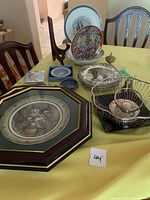 Overall view of the table with framed owl plate, Friendly Village plates set, metal wire baskets, and various additional plates and decorative items.