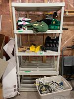 Five-tier resin shelf with assorted items on four shelves; bottom shelf contains plastic bin with cables and power strips.