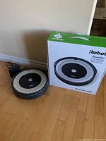 Roomba unit positioned next to its original box on hardwood floor