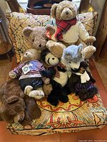 Group of seven stuffed animal plush toys including bears with tags and various clothing placed on a chair.