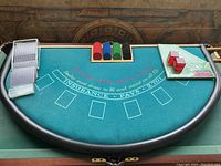 Photo of blackjack table showing playing cards, poker chips in four colors, and red dice on the green felt surface with printed game instructions.