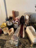 Overview of assorted candles, wax warmer, seashell in glass, taper candles, pine cone, and a figurine.