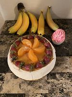 Photo showing a display of four fake bananas, a fake peach tart decorated with peach slices and strawberries, and a pink frosted fake cupcake on a marble surface.