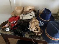 Multiple hats and belts arranged on a surface, including straw hats, knit hats, felt hats, and several belts in leather and fabric varieties.