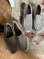 Two pairs of men's Skechers slip-on shoes, one pair dark brown and one pair light gray, placed on different surfaces including a carpet and wooden floor.